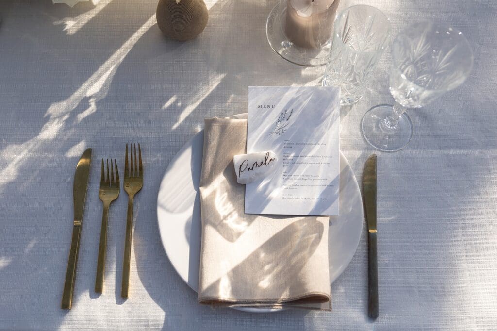 Table setting for a wedding with the name tag reading Pamela and a menu. White china and gold utensils with crystal glasses and set with white table linens.