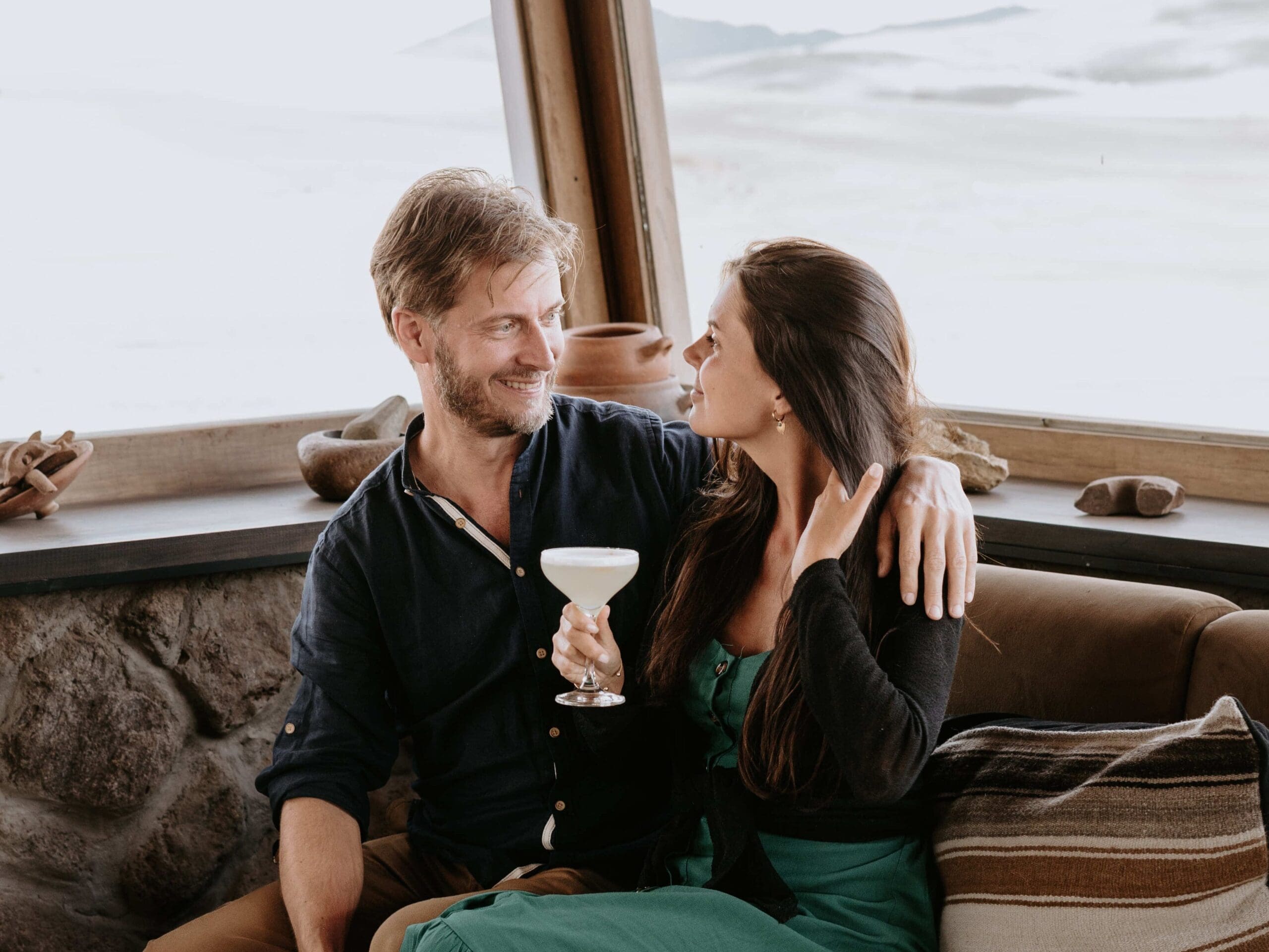 Photo of a couple with cocktails and sitting next to each other gazing in each others eyes. She has a cocktail.