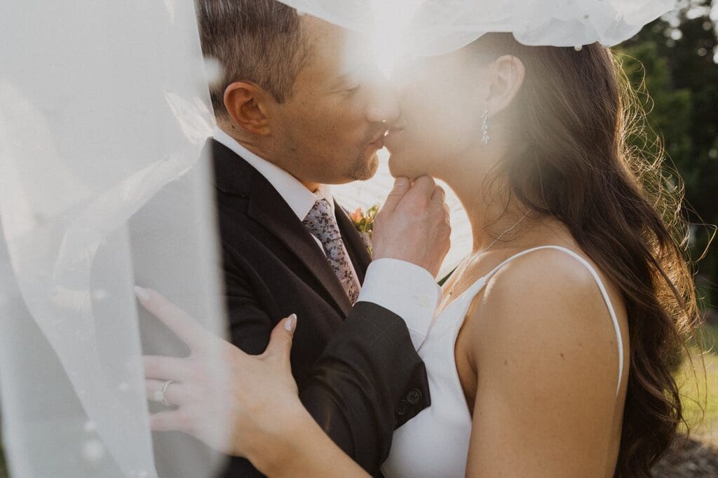 Man and woman under a wedding veil kissing with the sunlight shining through the veil. Celebrating their marriage.