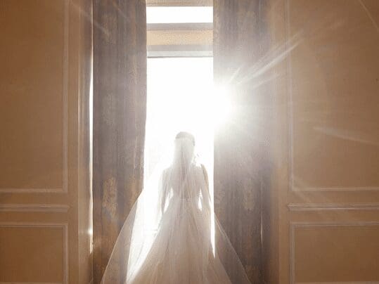 Photo of a Bride walking through the venue doors with the sunlight shining through her wedding veil.