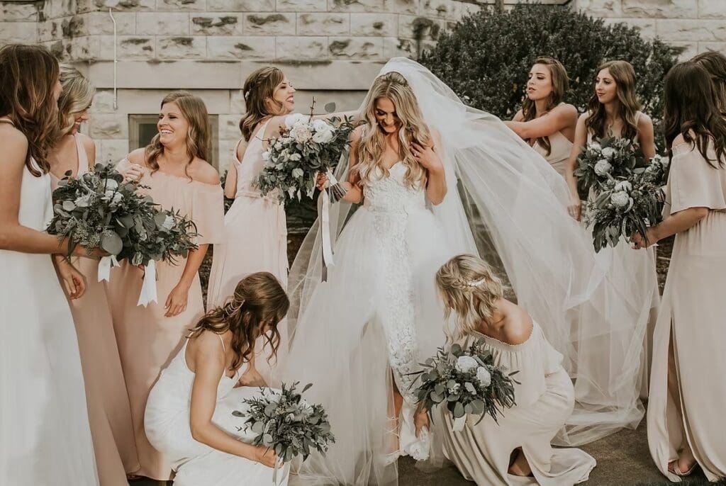 Bride with her bridesmaids have fun getting the bride ready for the ceremony against a white stone building. White flowers with greenery adorn the bride and bridesmaids. 