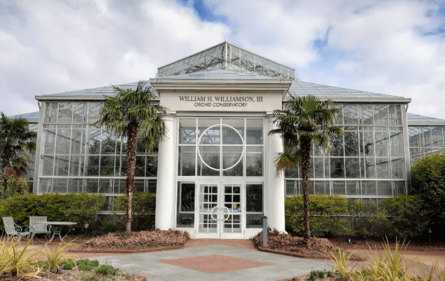 The facade of panes of glass atrium type building with palm trees and large white columns surrounded by lush gardens and walkways.
