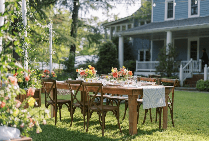 A Table setting with elegant china and lush tablescapes of flowers for guest to enjoy  on the grass setting, while surrounded by gardens and  the view of a farmhouse.