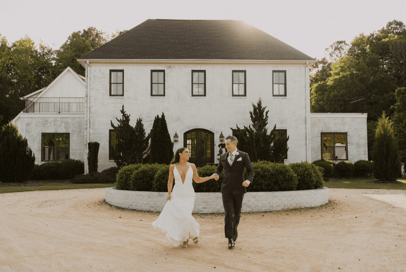 A bride and groom holding hands and running out in front of a white stone estate with lush greenery surrounding the home.