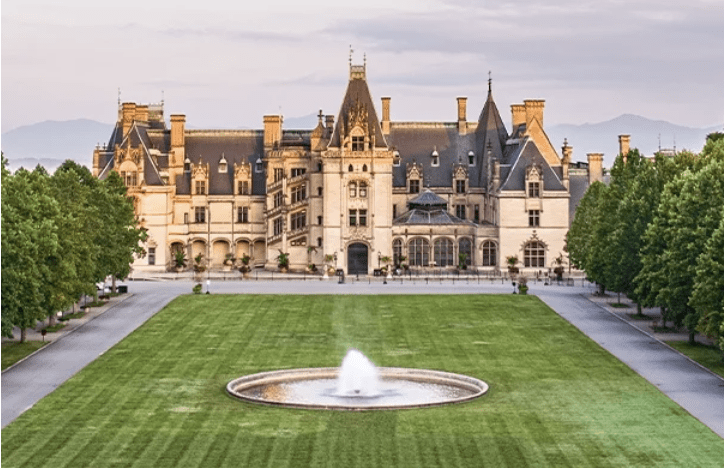 Front facade of the Biltmore in Asheville. Castle type appearance with large pitch roofs surrounded by lush manicured gardens and lawn. and trees lining the large fountain in front lawn.