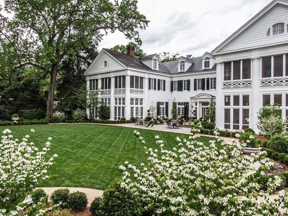 Photo of Duke Mansion stands tall with white columns and a grand portico. Green shutters frame the large windows. Lush Gardens and brick path.