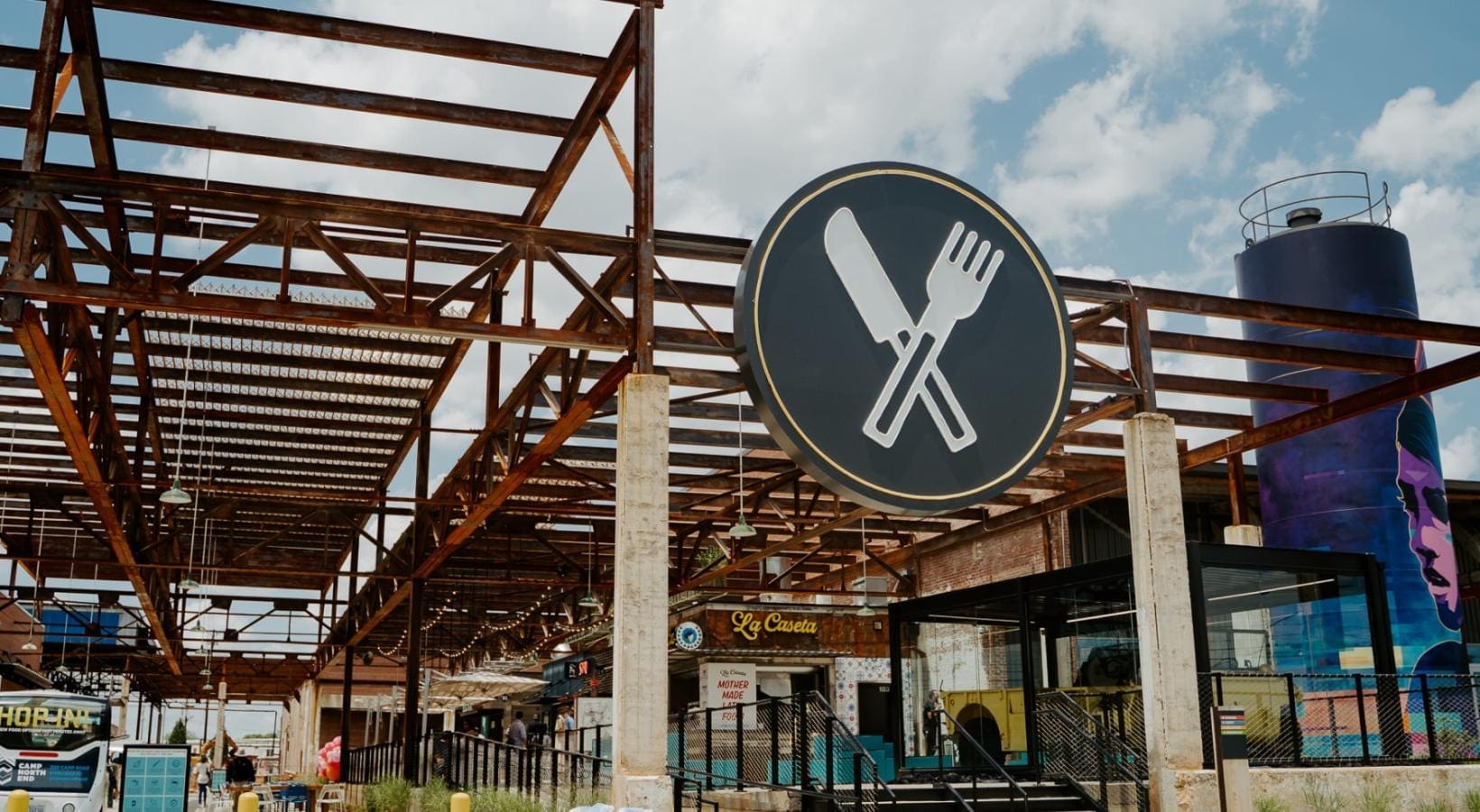 Camp North End facade with exposed metal beams and string lights with a huge logo of a knife and fork mounted on the front of the warehouse.