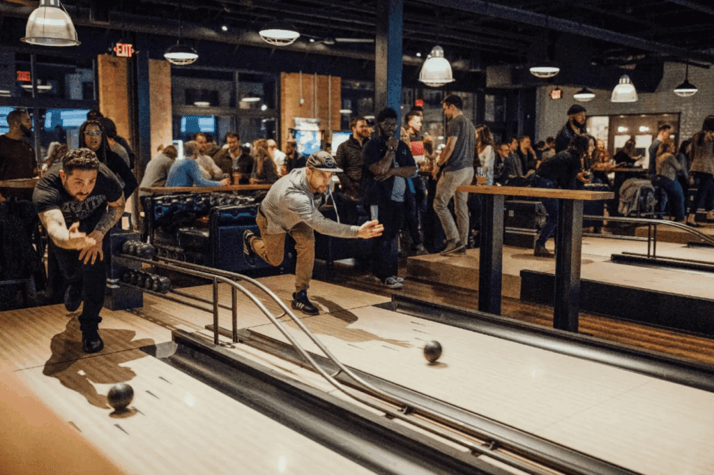 Duckpin bowlers at Pin Mechanical with hands extended and throwing the ball down the lane. A group of people at tables behind with food and drinks.
