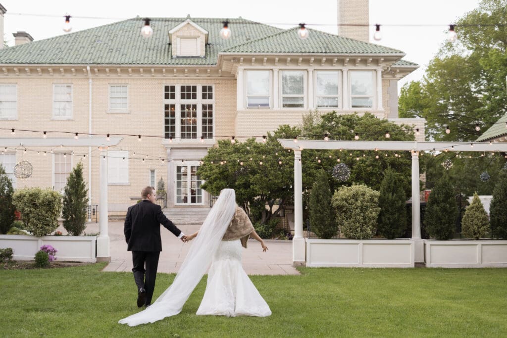 A bride and groom walking hand at  in hand at Separk Mansion in Charlotte NC wedding venue across a lush green lawn with string lights and garden landscaping. 