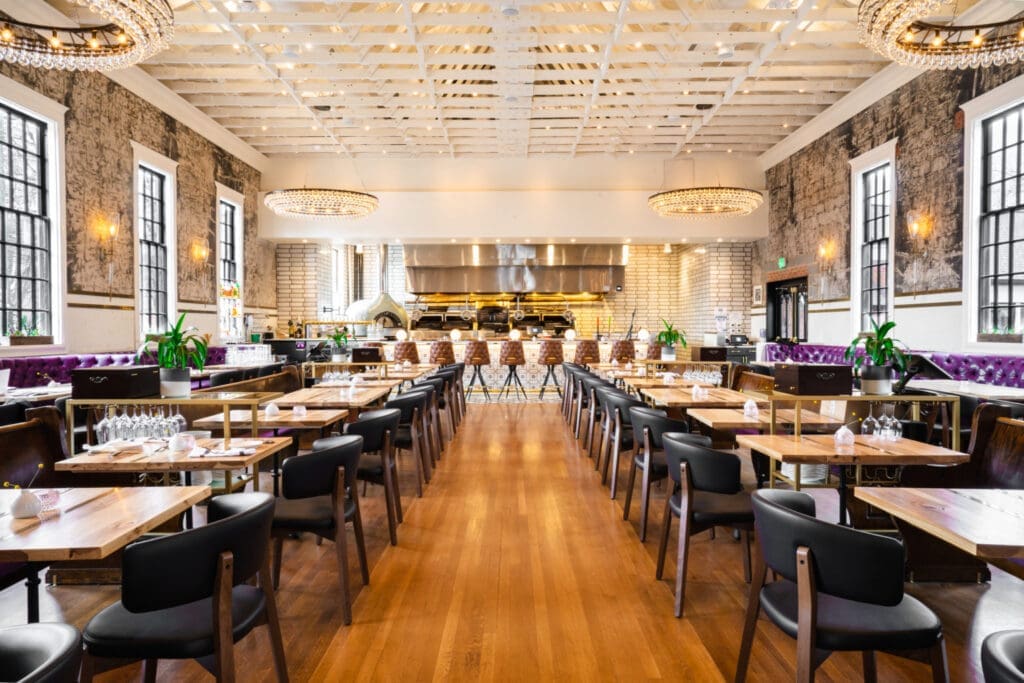 Supperland interior with wood floors and tables lining the walkways with a kitchen and bar at the end of the restaurant. Exposed beam ceiling with cylinder shaped light mounted on the ceiling.