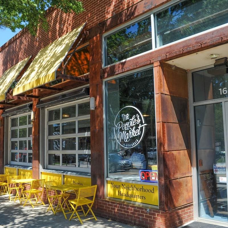 The Peoples Market Exterior with red orange bricks and yellow tables and chairs that line the garage door paned glass windows.