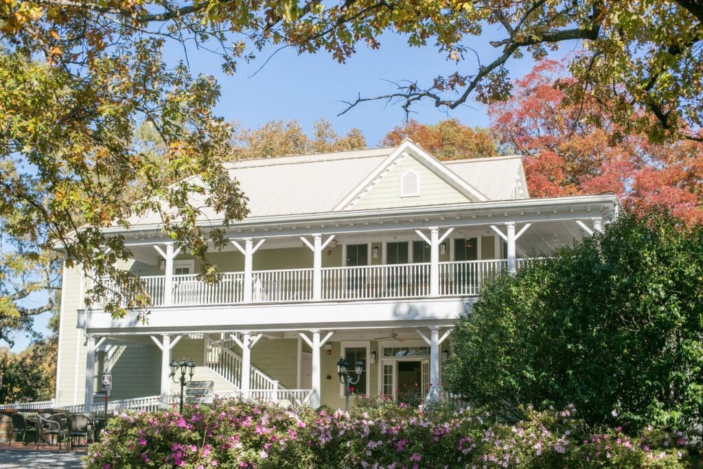 Whitehead Manor nestled in lush gardens with a wraparound porch with white trim and post facade with a sitting area for guest.